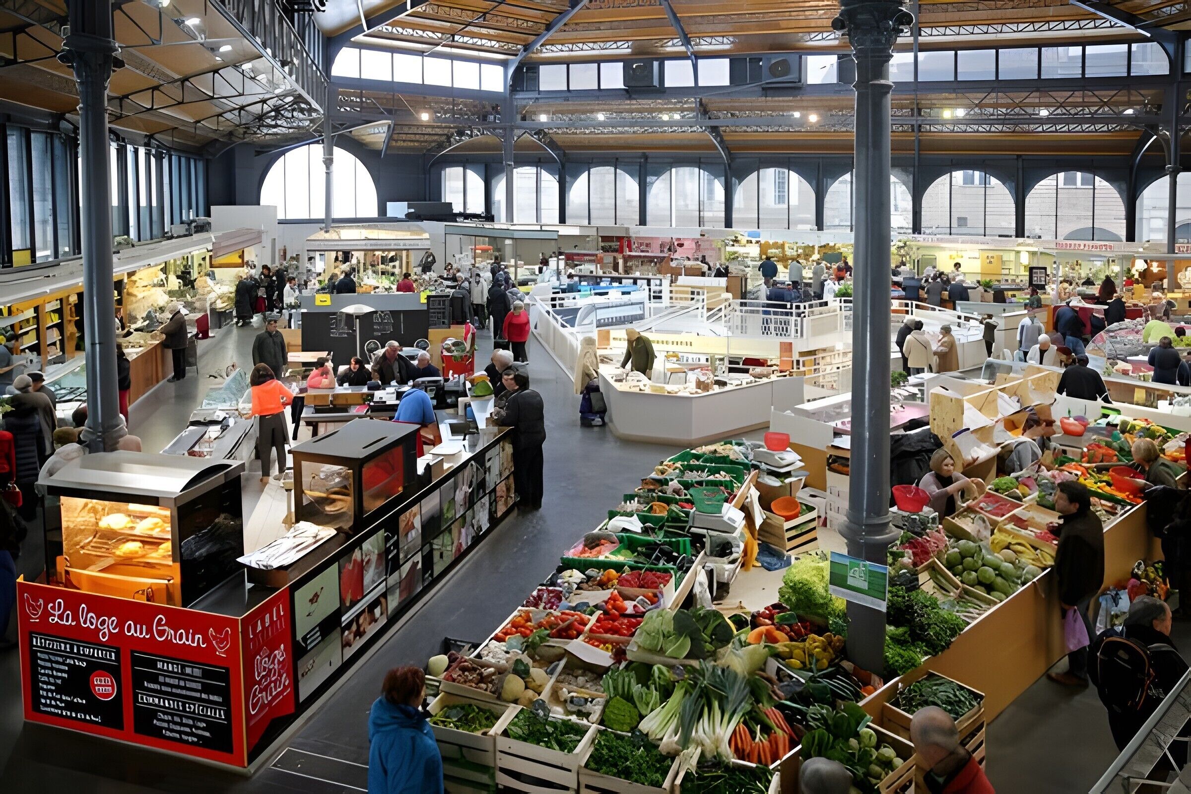 An indoor market with various stalls selling fresh produce and meats, surrounded by high ceilings and large windows.