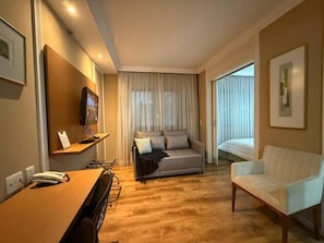 A hotel room with a wooden floor, a sofa, a chair, a desk with a phone, and a TV. - The Universe Executive Flat (São Paulo)