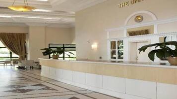 Een hotel lobby met een receptiebalie, een grote potplant en een bord met de tekst 'THE UNIVERSE'.