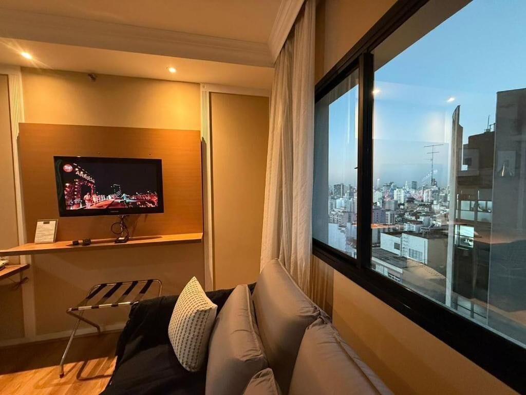 Ein modernes Hotelzimmer mit großem Fenster, einem an einer Holzablage montierten Flachbildfernseher, einer gepolsterten Sitzecke und Blick auf die Stadt.