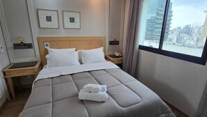 Una habitación de hotel con una cama grande, dos mesitas de noche, un ventanal con vistas a la ciudad y cuadros enmarcados en la pared.