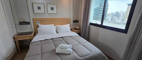 A hotel room with a large bed, two bedside tables, a window with a city view, and framed pictures on the wall.