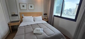 A hotel room with a large bed, two bedside tables, a window with a city view, and framed pictures on the wall. - The Universe Executive Flat (São Paulo)