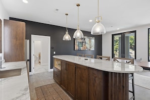 A modern kitchen with a marble countertop, wooden cabinets, pendant lighting, and a view to the outdoors.