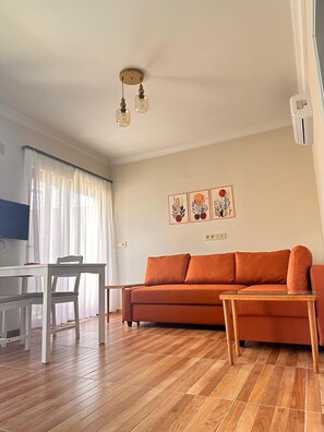 A living room with a wooden floor, a white table, a sofa, and a TV.