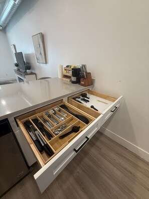 A kitchen with a white countertop, a drawer organizer with utensils, a coffee maker, and a wall-mounted shelf with plants and a picture.