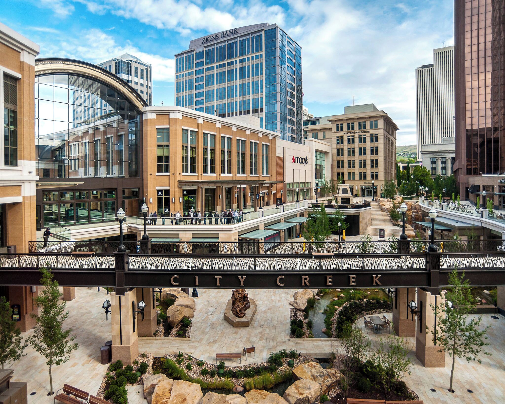 A multi-level shopping center with a prominent City Creek sign, modern buildings, and a landscaped plaza with seating areas.