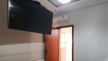 Quarto com uma cama, televisão na parede, ar condicionado e porta que dá para outro espaço.