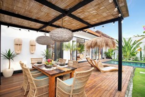 An outdoor dining area with a wooden table and wicker chairs, surrounded by a thatched roof and string lights. - 3 Bedroom Luxury Villa Blue Mango Umalas Canggu (Badung Regency)