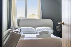 Ein ordentlich bezogenes Bett mit Kissen und einem gefalteten Handtuch, eine Nachttischlampe und ein Fenster mit Vorhängen.