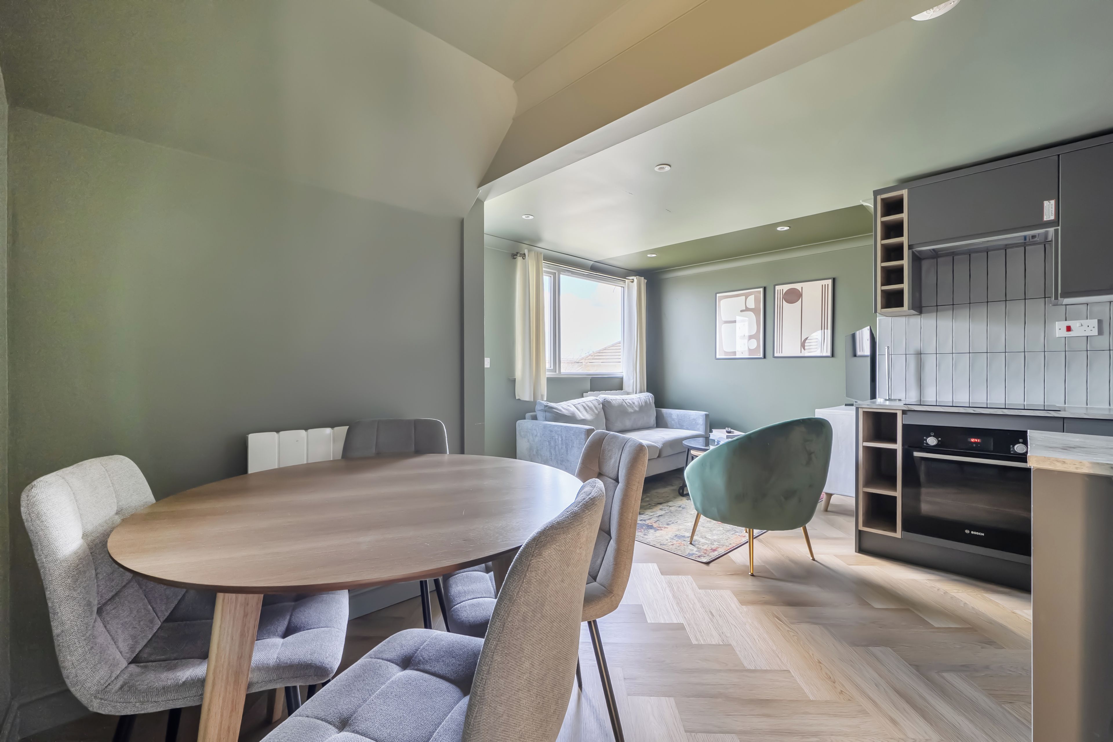 Eine moderne Küche mit einem Bosch-Ofen, eine Essecke mit einem Holztisch und grau gepolsterten Stühlen sowie ein graues Sofa in einem Wohnbereich mit Blick ins Freie.
