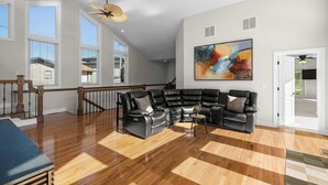 Ein geräumiges Wohnzimmer mit Ecksofa, Holzdielenboden, großen Fenstern und Deckenventilatoren.