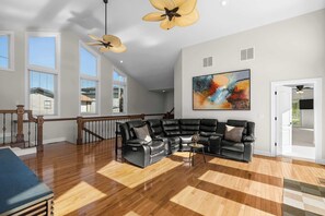 A spacious living room with a sectional sofa, hardwood flooring, large windows, and ceiling fans. - Sunrise Shores Estate - 4bd-4ba Luxury Lakefront (Camdenton)
