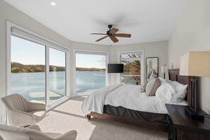 A bedroom with a large bed, a ceiling fan, a TV, and a view of a body of water. - Sunrise Shores Estate - 4bd-4ba Luxury Lakefront (Camdenton)