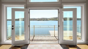 Ein Zimmer mit großen Fenstern, einer Holzdielen-Terrasse und Blick auf einen See sowie entfernte Gebäude.
