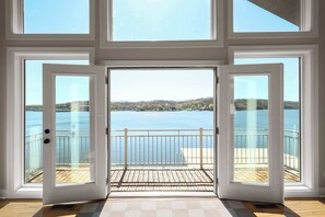 A room with large windows, a wooden deck, and a view of a lake and distant buildings. - Sunrise Shores Estate - 4bd-4ba Luxury Lakefront (Camdenton)