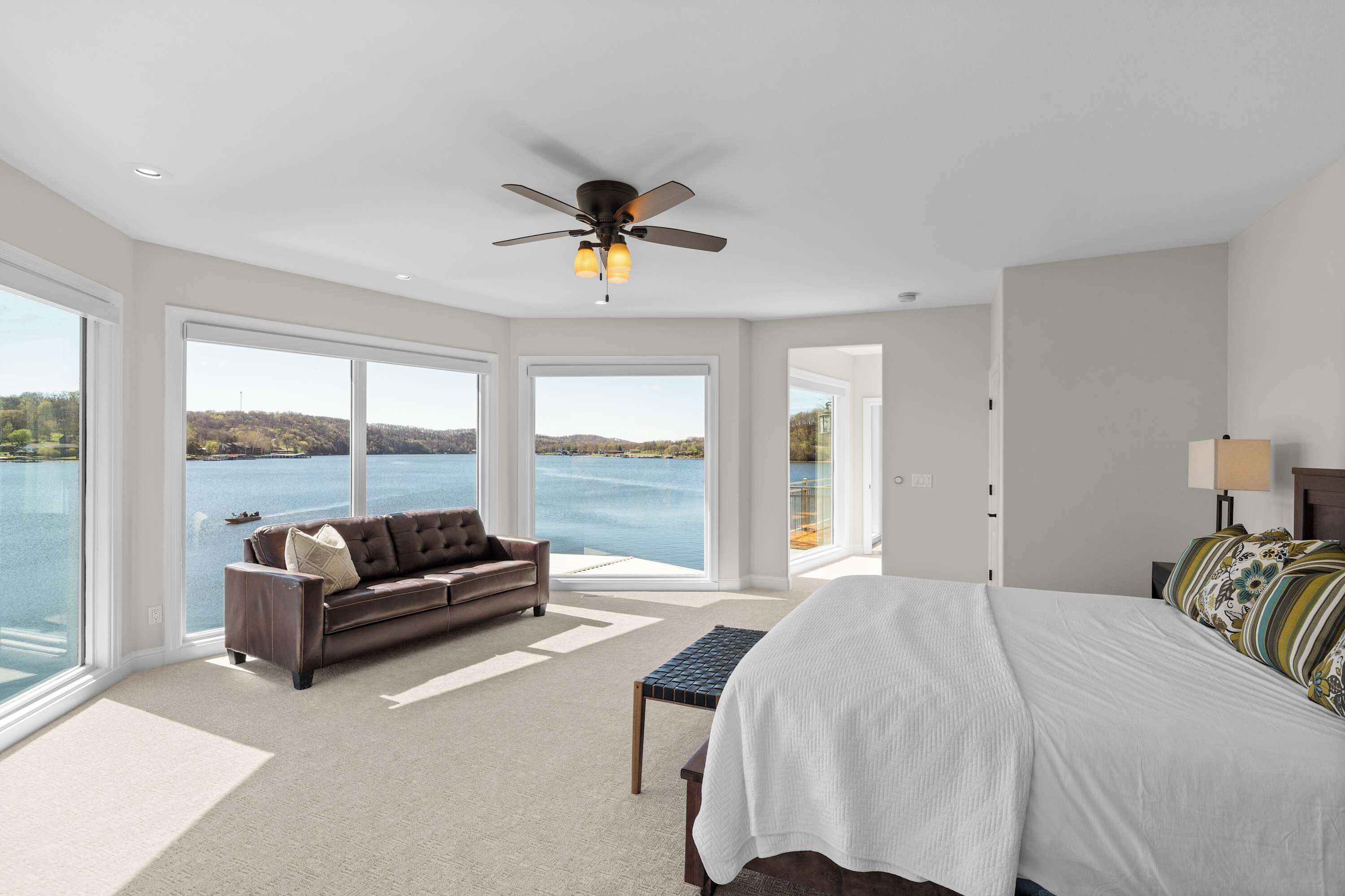 A bedroom with a bed, a sofa, a ceiling fan, and large windows overlooking a body of water.