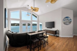 A modern living room with a large window, a flat-screen TV, and a dining area with a table and chairs. - Sunrise Shores Estate - 4bd-4ba Luxury Lakefront (Camdenton)
