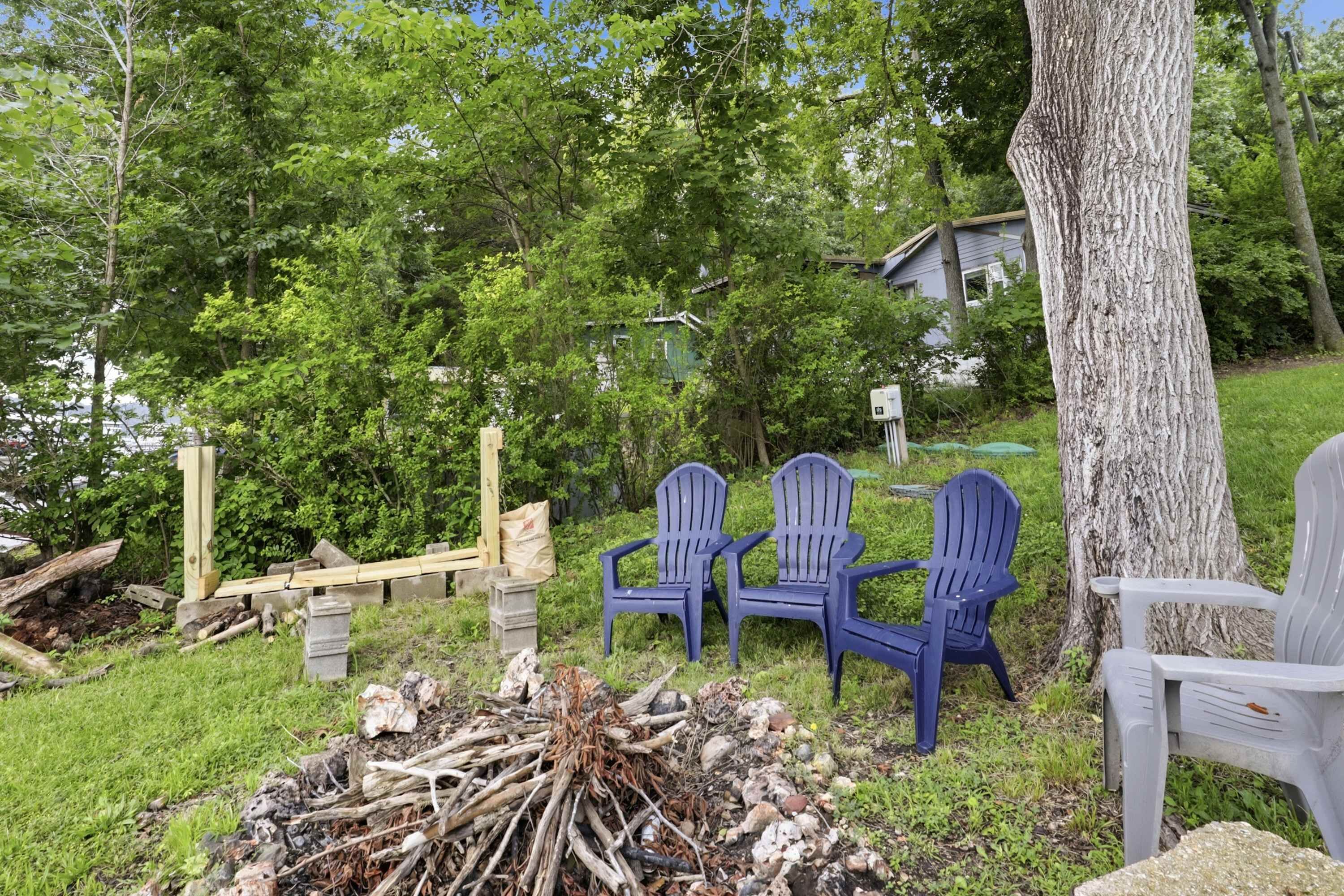 A backyard with a pile of firewood, two blue Adirondack chairs, and a tree.