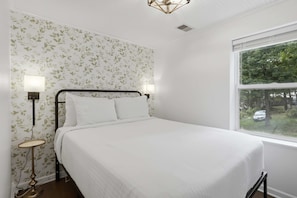 A bedroom with a bed, two bedside lamps, a window with a view of greenery, and a patterned wallpaper. - Visit This 4 Bedroom - 3 Bath Lakefront Home With Private Dock (Stover)