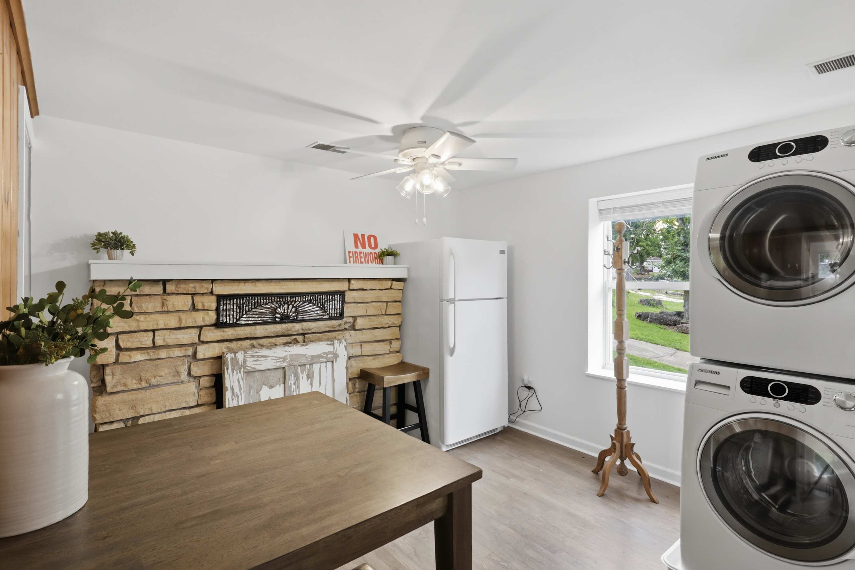 A laundry room with stacked washers and dryers, a ceiling fan, a refrigerator, and a window with a view of greenery.