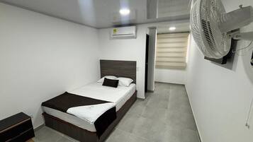 Een slaapkamer met een bed, nachtkastje, een wandairconditioner en een staande ventilator.