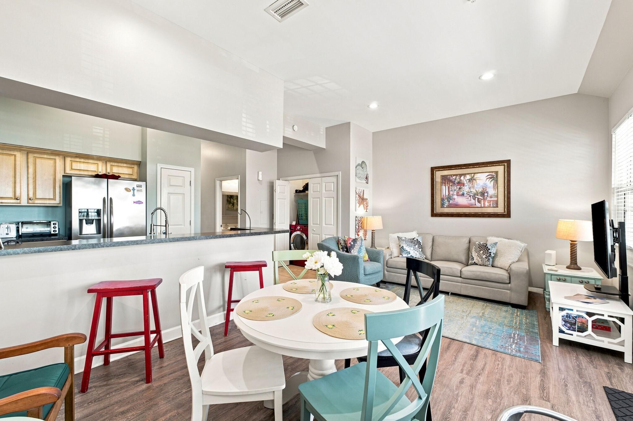 A modern kitchen with a built-in oven, a dining area with a round table and chairs, a living room with a sofa and a TV, and a laundry room visible through an open door.