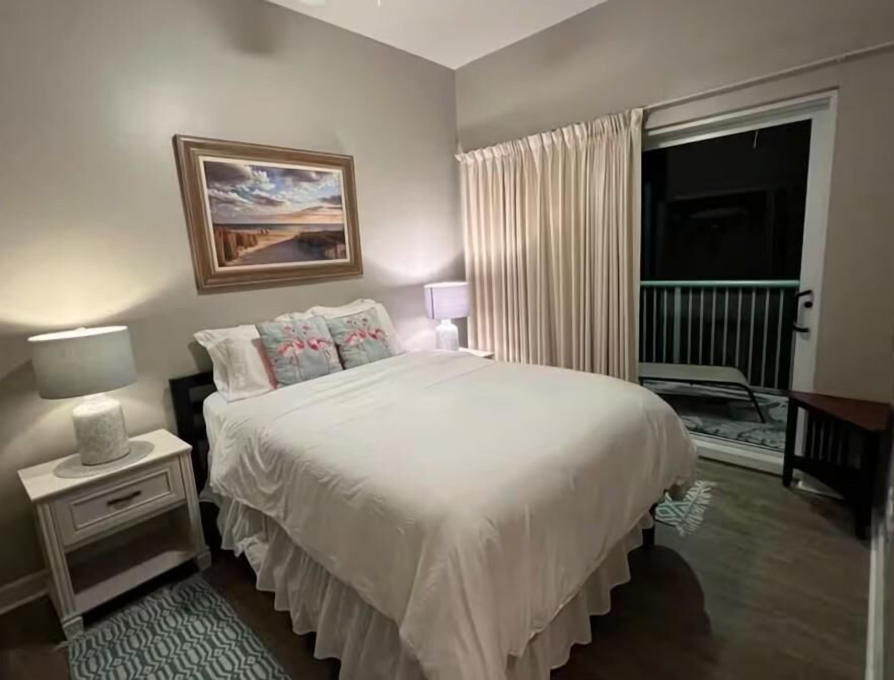 A bedroom with a bed, nightstands, a lamp, a painting, a window with curtains, and a balcony door.