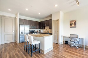 A modern kitchen with dark cabinets, a stainless steel refrigerator, a built-in oven, and a white island with bar stools. - Downtown Houston 1BR • Balcony • Fast Wi-Fi • Free In-Unit Laundry (Houston)