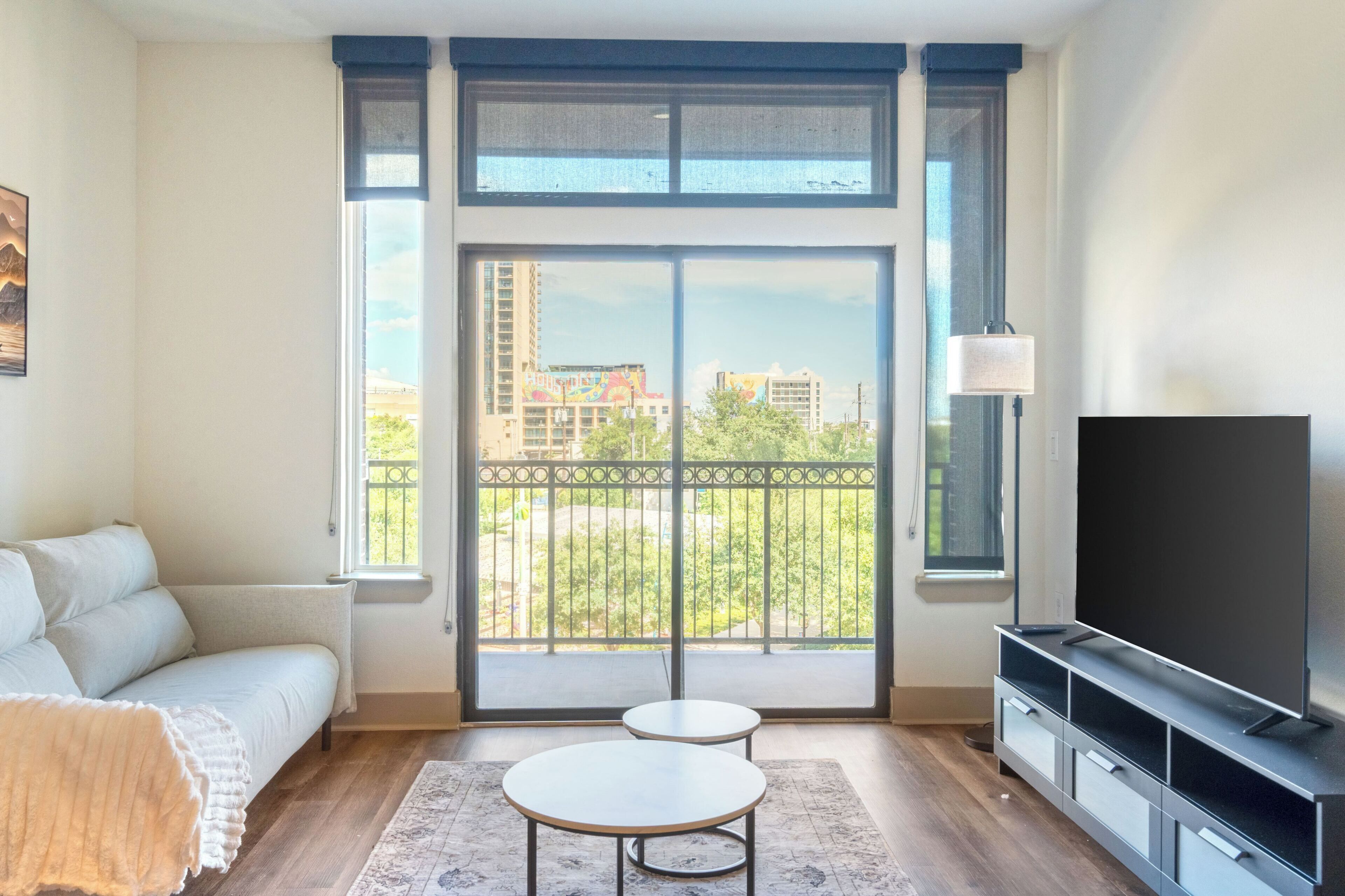 A modern living room with a sofa, a TV, a small table, and a balcony with a view of buildings and greenery.