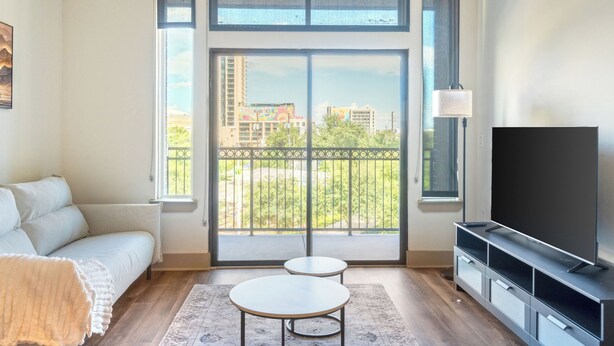 A modern living room with a sofa, a TV, a small table, and a balcony with a view of buildings and greenery. - Downtown Houston 1BR • Balcony • Fast Wi-Fi • Free In-Unit Laundry (Houston)