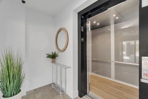 Un interior moderno con una mesa consola de vidrio transparente, una planta en maceta, un adorno de pared tejido y una puerta de ascensor de vidrio.