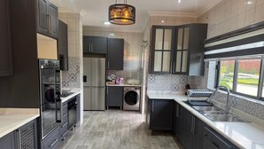Eine moderne Küche mit dunklen Schränken, einem Edelstahl-Kühlschrank, einer Waschmaschine und einem Fenster mit Blick ins Freie.