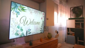 A modern living room with a large flat-screen TV displaying 'Welcome' on a floral background, a wooden cabinet, a green vase, and a small table. - Cozy 1-bedroom studio in beautiful Marrakech with AC (Marrakech)