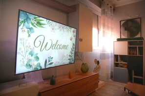 Ein modernes Wohnzimmer mit einem großen Flachbildfernseher, der 'Welcome' vor floralem Hintergrund zeigt, einem Holzschrank, einer grünen Vase und einem kleinen Tisch.