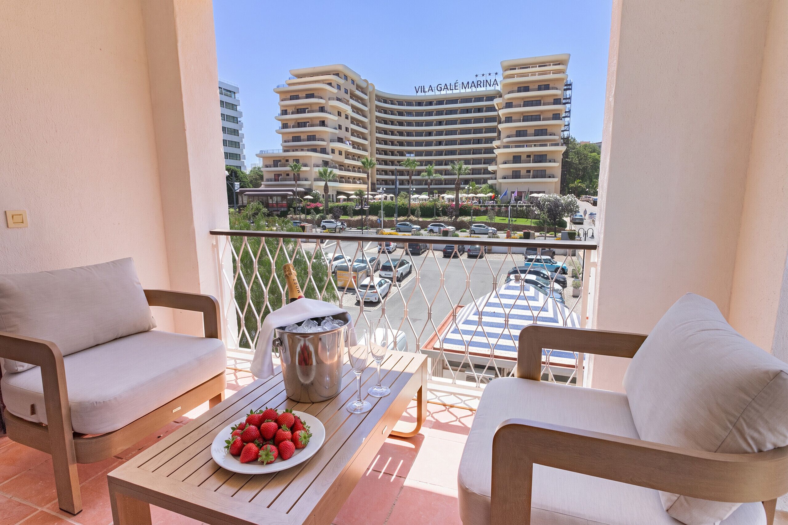 A balcony with a table, chairs, a plate of strawberries, a champagne bucket, and a view of a hotel named 'Vila Galé Marina' with a parking lot and palm trees.