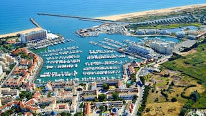 An aerial view of a coastal marina with numerous boats docked, a sandy beach, and a mix of residential and commercial buildings. - Apartamento Vintage | 4 bedrooms | Vilamoura Marina (Vilamoura)