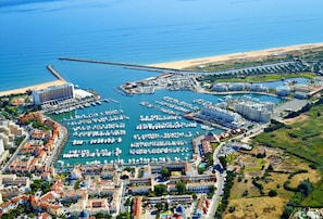 An aerial view of a coastal marina with numerous boats docked, a sandy beach, and a mix of residential and commercial buildings. - Apartamento Vintage | 4 bedrooms | Vilamoura Marina (Vilamoura)