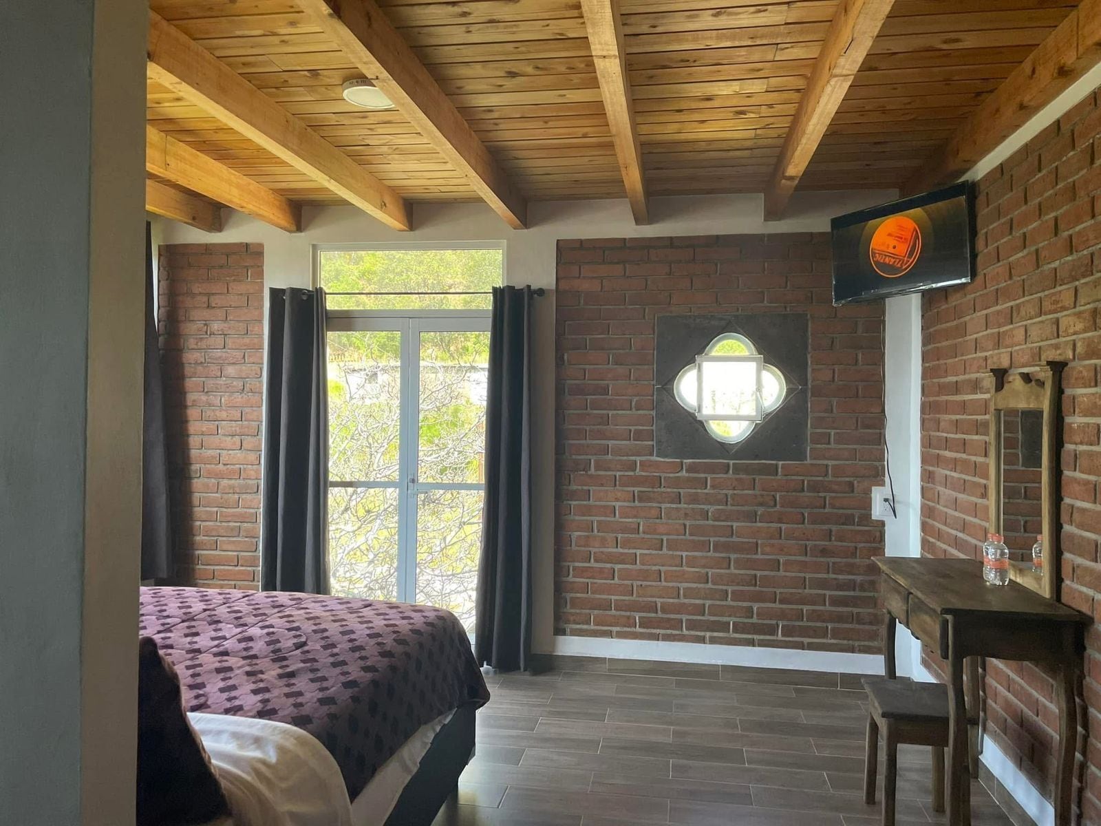 A room with a bed, a desk, a brick wall, and a window with curtains.