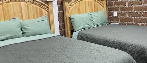 A room with two beds, each with a wooden headboard and green bedding. The walls are made of exposed brick.