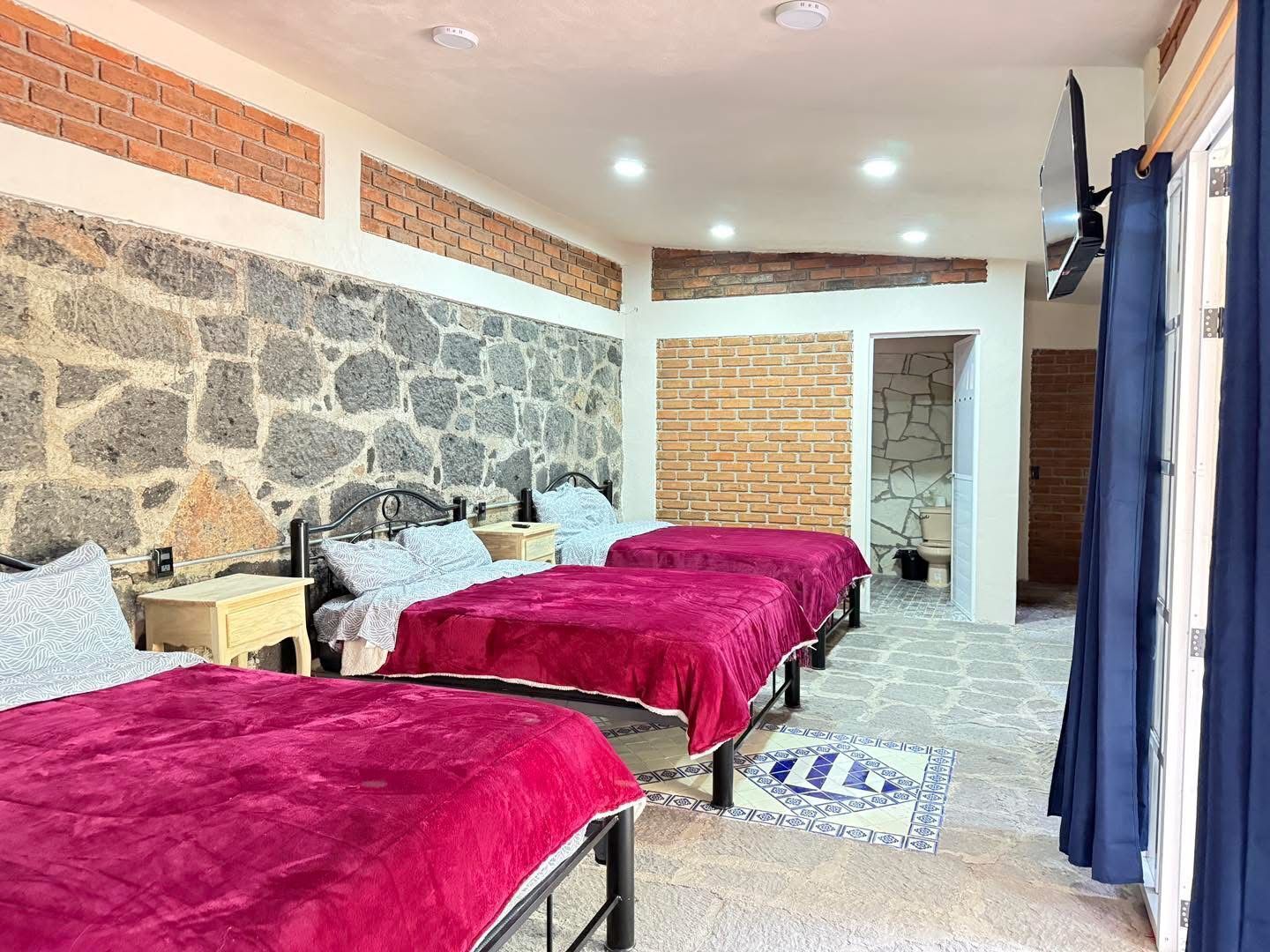 Una habitación con dos camas, paredes de piedra, piso de baldosas y un televisor fijado a la pared.