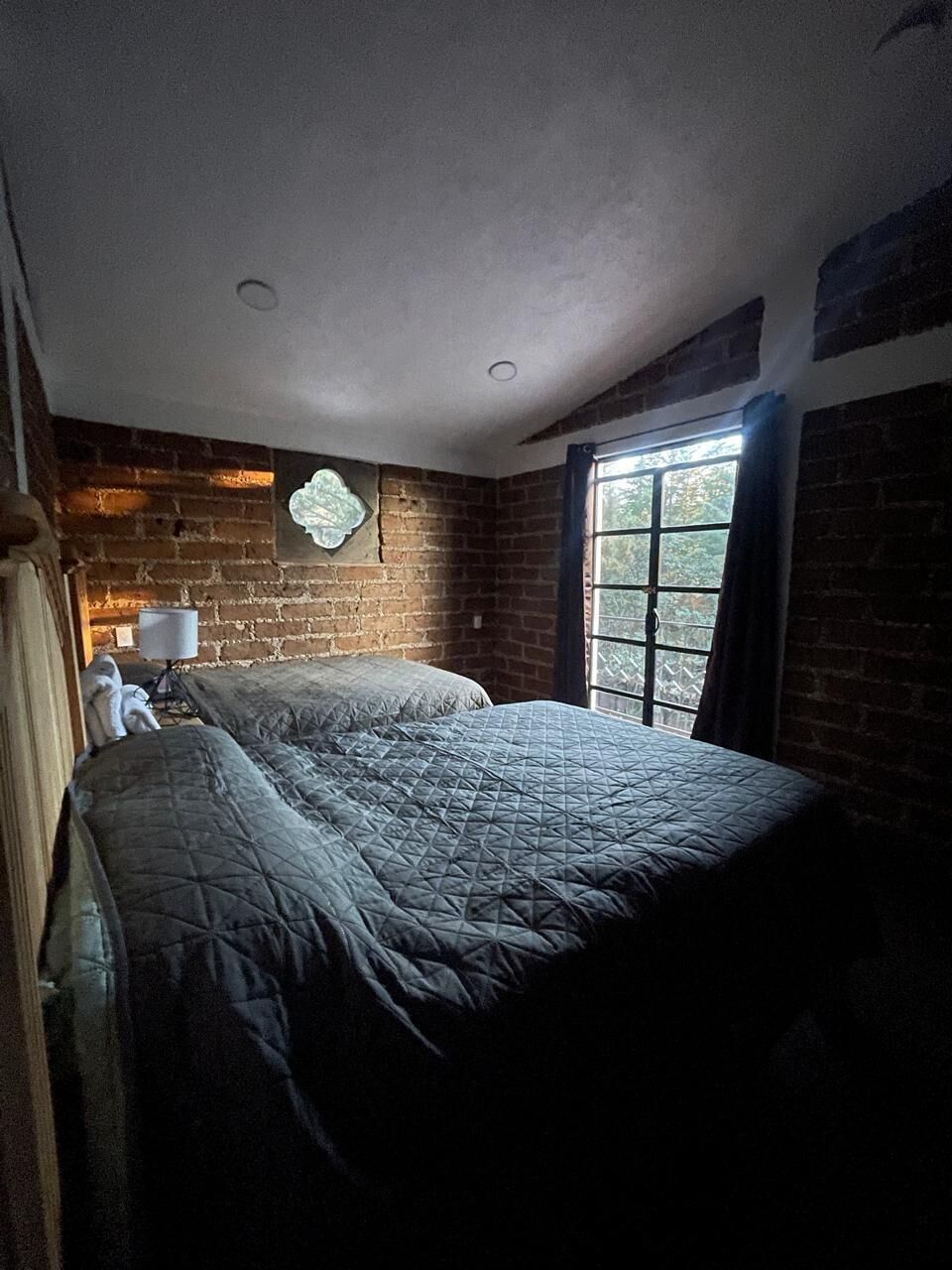Una habitación con pared de ladrillo, ventana con cortinas y cama con colcha acolchada.