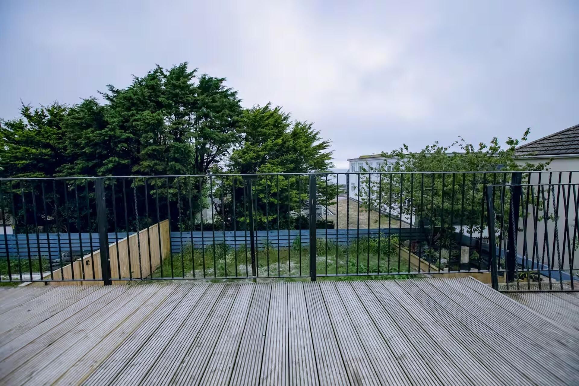 Eine Holzterrasse mit einem Metallzaun, Bäumen und einem Gebäude im Hintergrund.