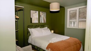 Un dormitorio con cama, una silla, mesita de noche, lámpara y ventana.