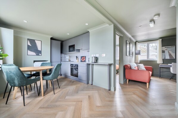 A modern kitchen with a built-in oven and microwave, a dining area with a wooden table and chairs, and a living area with a sofa and a window. - 1 Bedroom Apartment Sleeps 2 - Pets Welcome (St Ives)