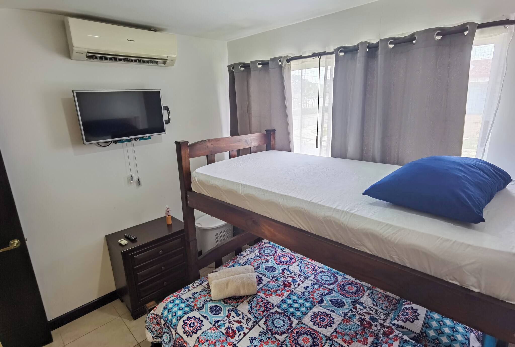 A bedroom with a bed, a TV mounted on the wall, an air conditioner, a window with curtains, and a small bedside table with a lamp. - Acogedora Casa de Playa, Condominio Malaga Bejuco (Parrita)