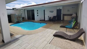 Piscina con borde de baldosas, una silla de reposo y una zona de cocina con armarios y lavamanos.