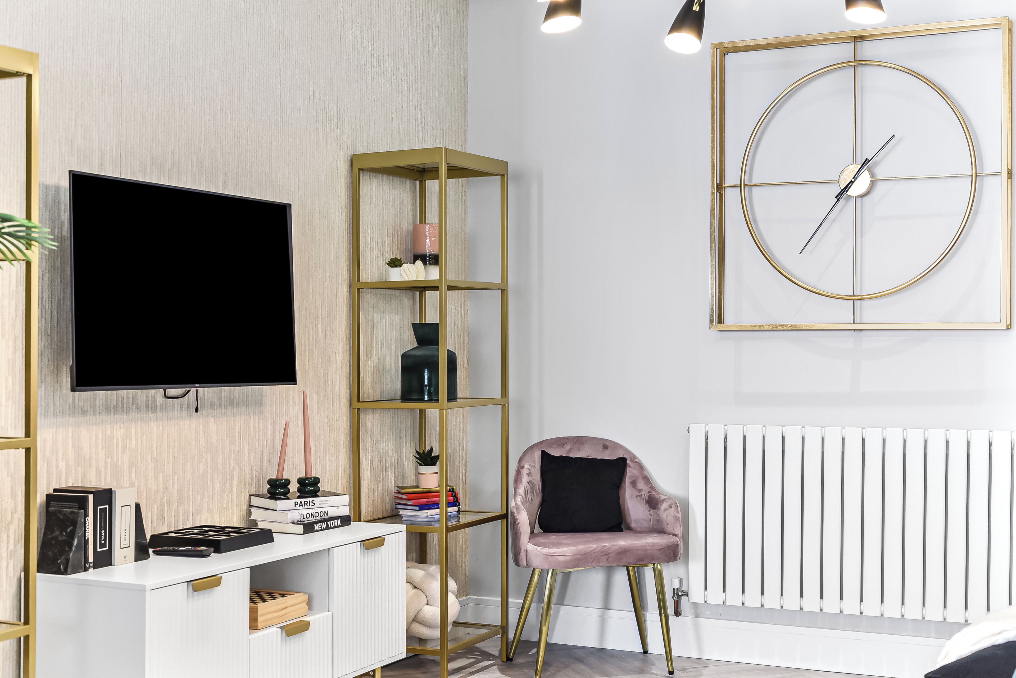 A modern living room with a flat-screen TV, a gold-framed bookshelf, a pink armchair, and a round wall clock.