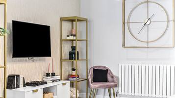 A modern living room with a flat-screen TV, a gold-framed bookshelf, a pink armchair, and a round wall clock.