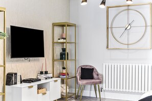 Ein modernes Wohnzimmer mit Fernseher, einem goldfarbenen Bücherregal, einem rosa Sessel und einer runden Wanduhr.